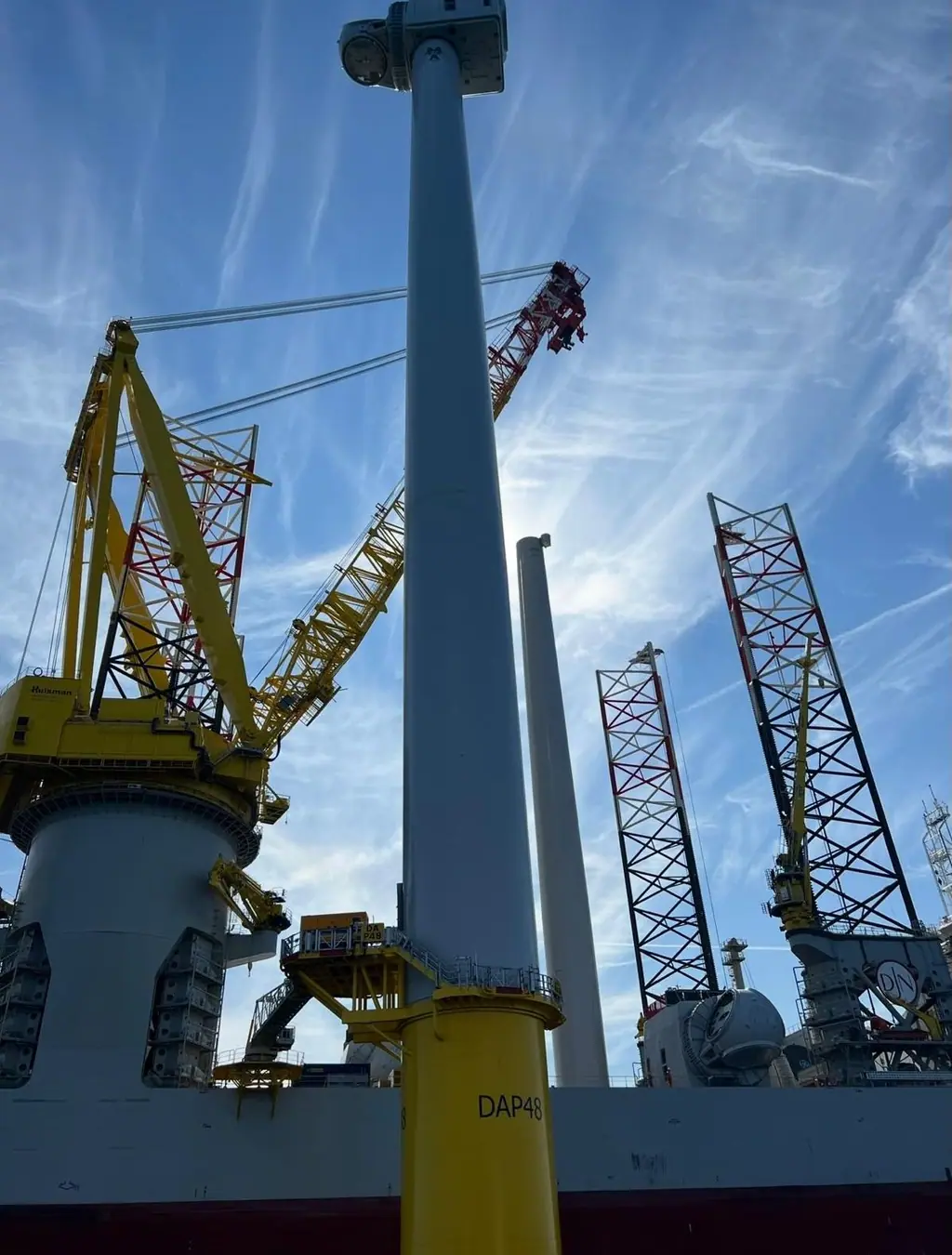 Offshore wind turbine tower on a yellow transition piece, seen from deck level beside the heavy-lift crane and jack-up legs of an installation vessel